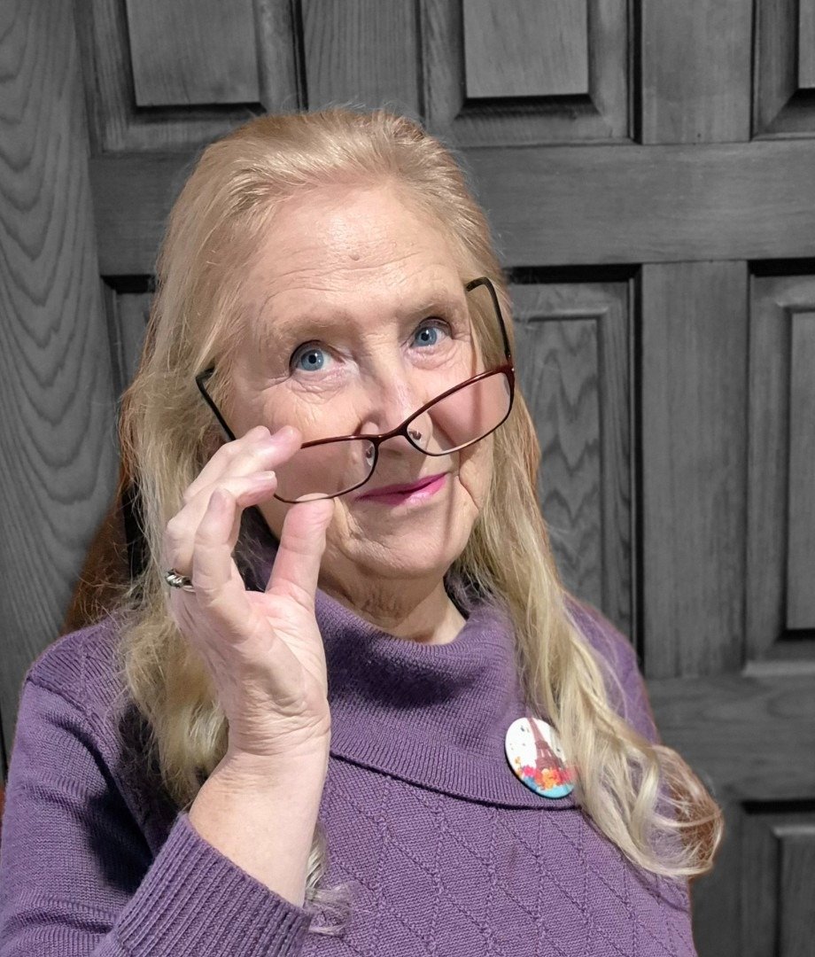 Joy Lubawy holding her glasses, wearing a purple jumper